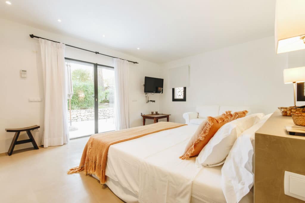 Bright bedroom perfect for luxury travel, featuring a large bed adorned with white linens and an orange throw. A small table and chair sit by a wall-mounted TV. Sliding glass doors on the left open to a garden area, enhancing the rooms charm with its cream-colored walls and floors. David Pearson Travel