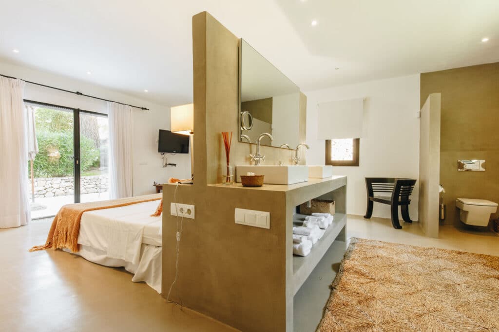 Experience luxury travel in this spacious hotel room featuring a large bed and orange accents. Natural light floods through a sliding glass door with sheer curtains, illuminating the elegant partitioned bathroom area. A textured rug complements the polished floor, with towels neatly arranged under the sink. David Pearson Travel