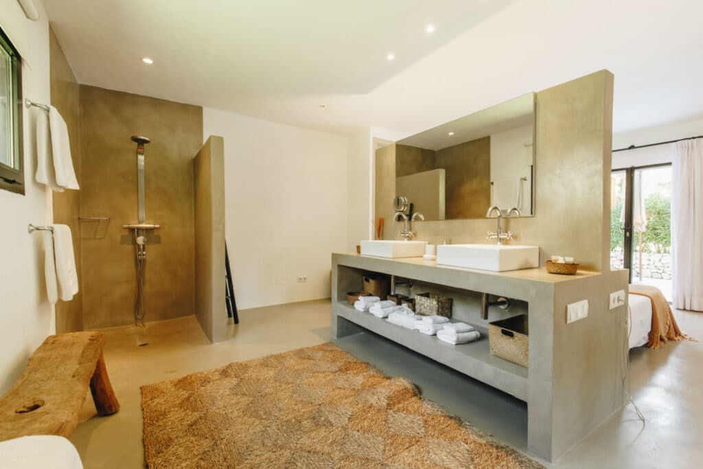 Spacious bathroom with a double sink vanity, wall mirrors, and open shelving stocked with towels and baskets. A large walk-in shower graces one corner while a textured rug adorns the floor, all basking in natural light from the window—blending homey comfort with the feel of luxury travel. David Pearson Travel
