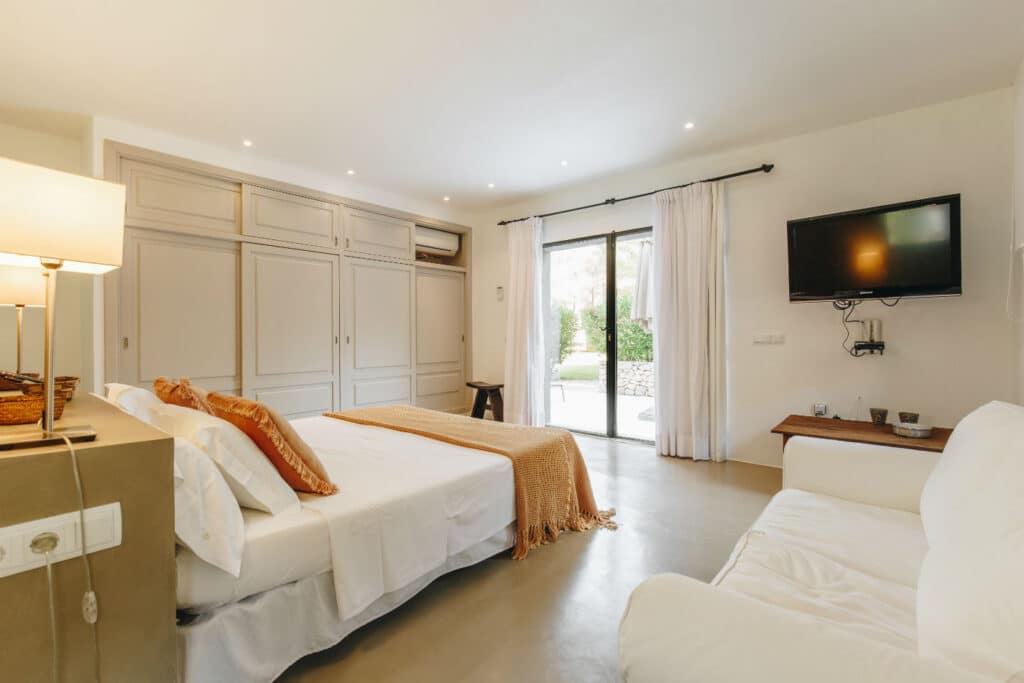 Experience luxury travel in this cozy bedroom with white walls, featuring a neatly made bed with orange accents, a wall-mounted TV, and a large wardrobe. Curtains frame a sliding glass door leading to lush greenery. A sofa near the TV and soft lighting complete the warm ambiance. David Pearson Travel