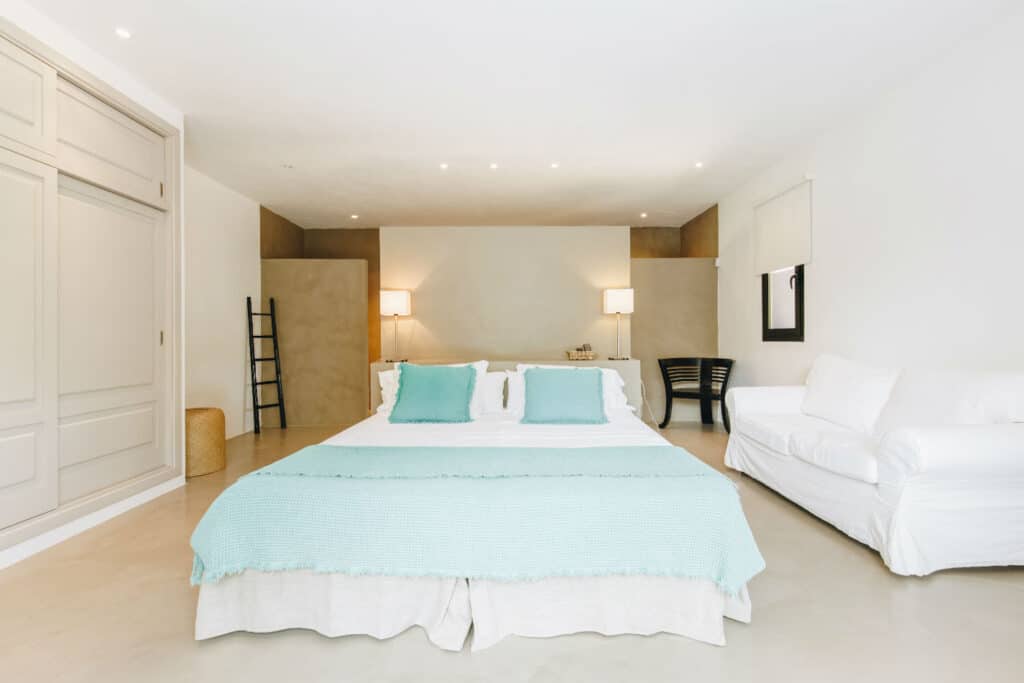 A minimalist bedroom evokes the serenity of luxury travel, featuring a large bed with turquoise pillows and a light blue blanket. On either side, there are lamps and small tables. A white sofa sits to the right, and a black chair near the window. The walls and floors are light-colored. David Pearson Travel