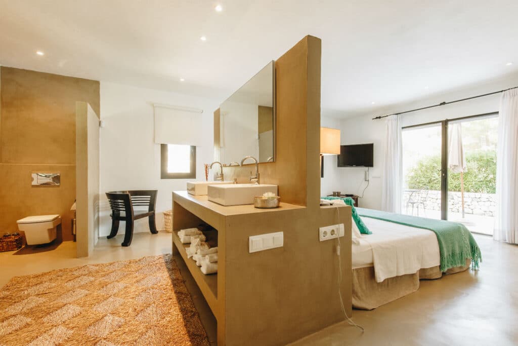 A modern bedroom with a spacious, open layout exuding a luxury travel vibe. A double bed with a green throw blanket faces a large glass door, inviting the outside in. A partition with a sink and mirror separates the bed from the toilet area. White walls and natural light enhance the bright atmosphere. David Pearson Travel
