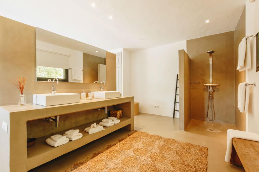 Spacious modern bathroom, perfect for luxury travel, features a large mirror above a dual sink vanity. Towels are neatly arranged on shelves below. A walk-in shower is on the right with a ladder-style towel rack beside it. The room is elegantly decorated in neutral tones. David Pearson Travel