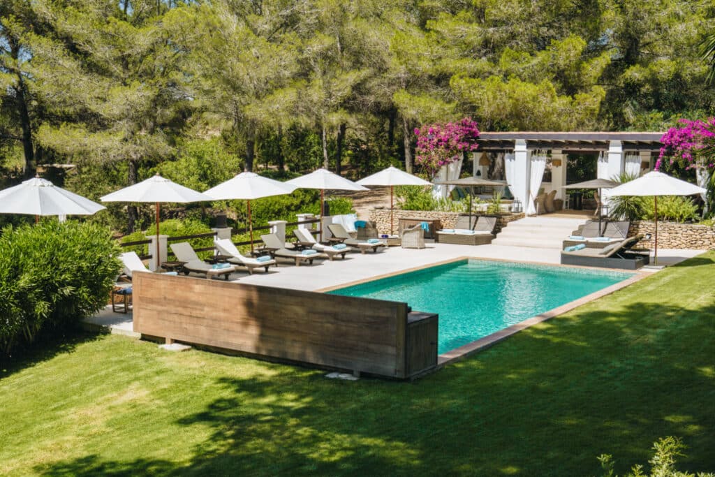 A luxurious outdoor pool area, perfect for luxury travel enthusiasts, is surrounded by sun loungers with white umbrellas. A shaded cabana with curtains and seating nestles among lush greenery and blooming bougainvillea, offering an idyllic escape. David Pearson Travel