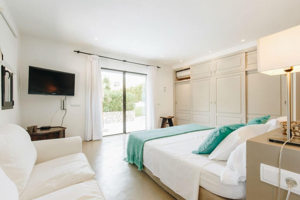 A bright bedroom perfect for luxury travel features a large bed adorned with white sheets and teal accents. A sofa sits on the left, opposite a wall-mounted TV and a spacious white wardrobe. Sliding glass doors open to a garden, while soft lighting enhances the clean, modern decor. David Pearson Travel