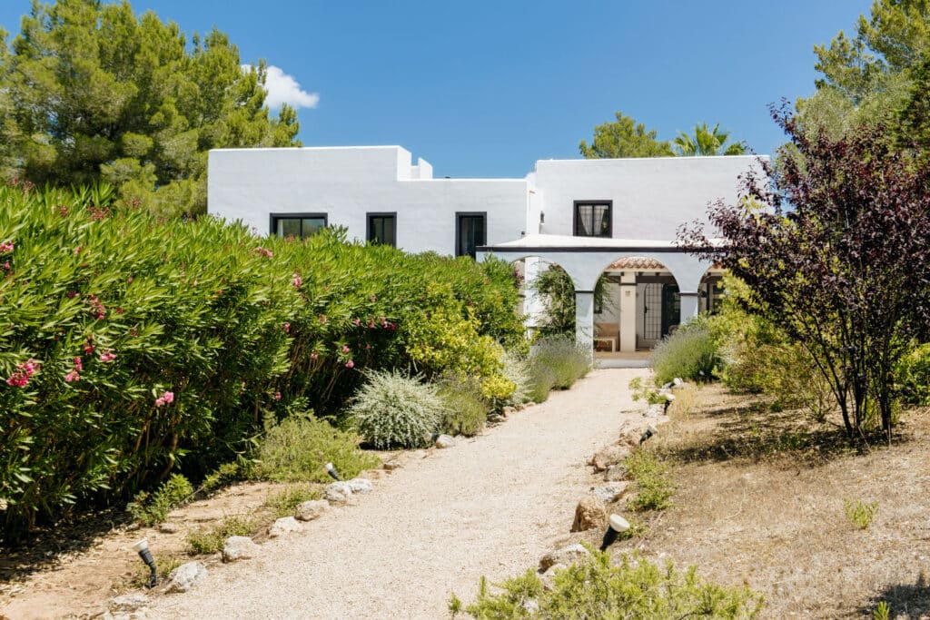 A Mediterranean-style white house with flat roofs, embraced by lush greenery and bushes, offers a luxury travel escape. A gravel pathway elegantly guides you to the entrance under a clear blue sky. David Pearson Travel