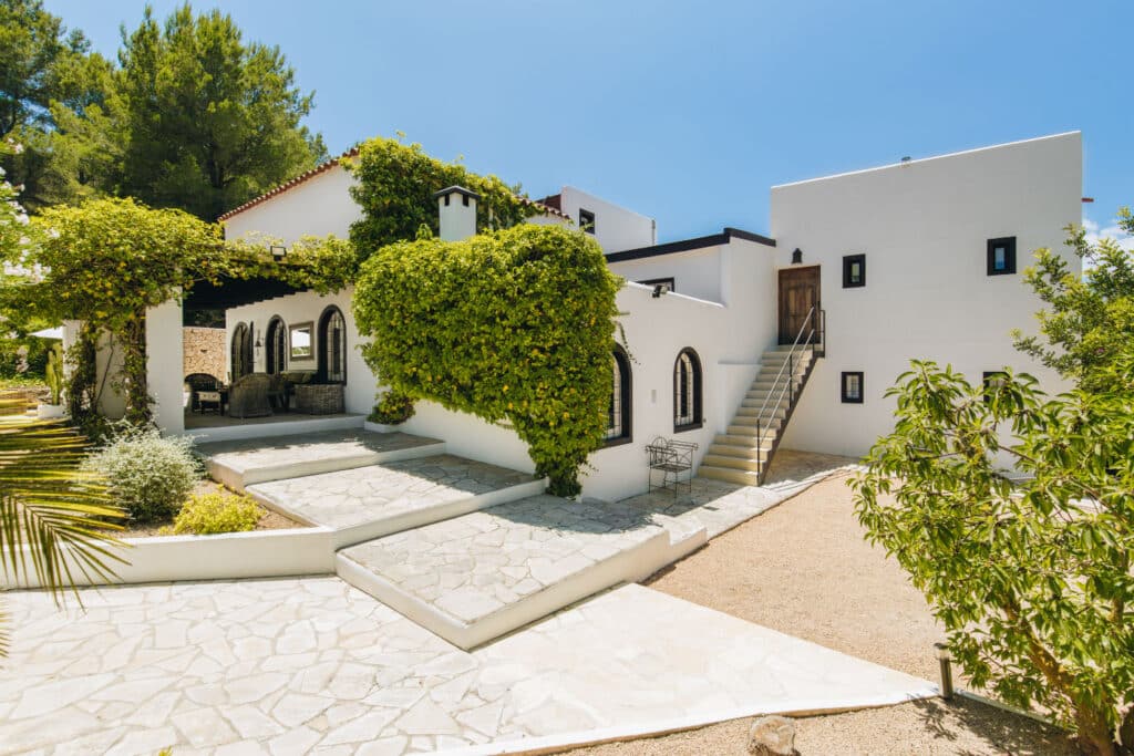 A white, modern Mediterranean-style house with arched windows, covered in green ivy, offers the perfect backdrop for luxury travel. It features a stone walkway and a staircase leading to an upper-level door, set against sparse vegetation and a sunny, clear blue sky overhead. David Pearson Travel
