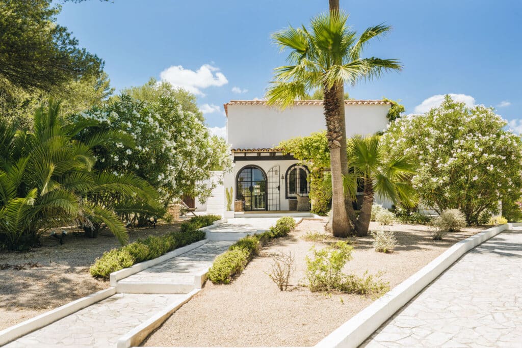 A white stucco house with a tile roof, embodying luxury travel, is surrounded by lush greenery, including palm and flowering trees. A stone path leads to the entrance, and the sky is clear and blue. David Pearson Travel