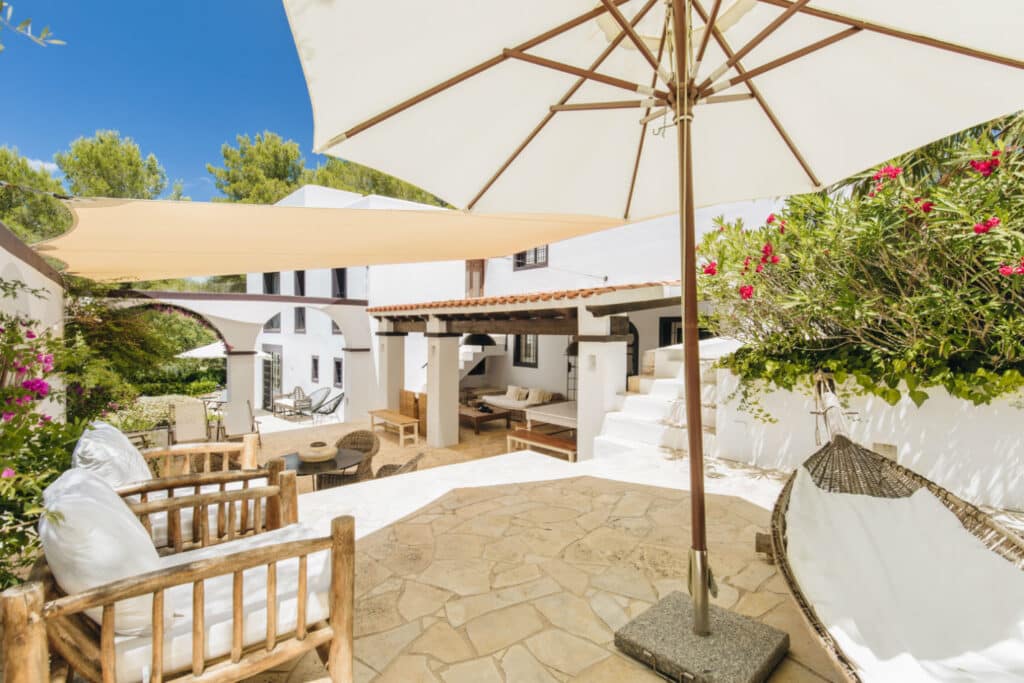 A sunny outdoor patio with white walls and rustic wooden furniture offers a touch of luxury travel. White cushions adorn the chairs beneath a large beige parasol, while a staircase leads to an elegant terrace. Greenery and pink flowers add vibrancy to this bright, inviting space. David Pearson Travel