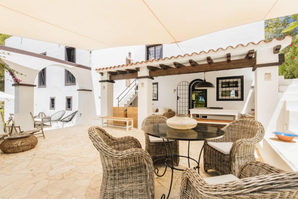 Experience luxury travel right at home on this outdoor patio, where wicker chairs and a round glass table sit beneath a covered terrace. Adorned with white walls, stone flooring, and elegant hanging lights, the area invites you to unwind with its plush sofa and decorative vases. David Pearson Travel