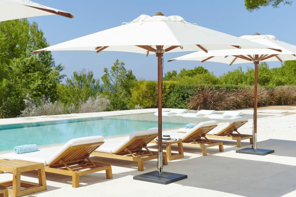 A row of wooden lounge chairs with white cushions and blue towels is elegantly arranged by a rectangular pool. White umbrellas provide shade, epitomizing luxury travel. Lush green trees and a clear blue sky form the backdrop, creating a serene and relaxing atmosphere. David Pearson Travel