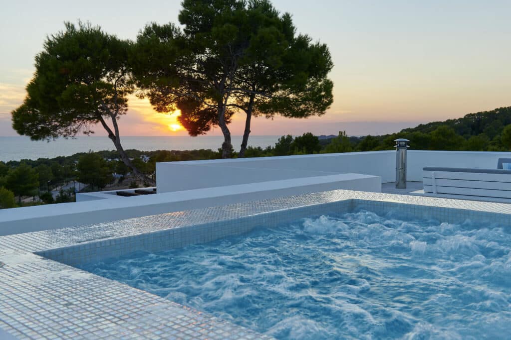 Experience luxury travel at its finest with a bubbling hot tub on a terrace, complete with a nearby bench. Overlook the scenic landscape as the sun sets behind silhouette trees, casting a warm glow over distant hills and ocean views. David Pearson Travel