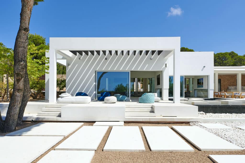This modern white house exudes luxury travel vibes with its sleek design featuring large glass doors and windows. The minimalist patio boasts white seating adorned with decorative pillows, while stone and pebble pathways guide you to the elegant entrance. A tree graces the left side for added charm. David Pearson Travel