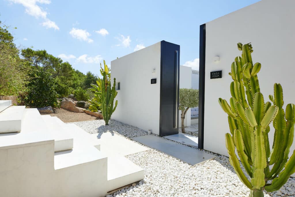 Modern white buildings with a minimalist design exude luxury travel vibes, surrounded by green cacti and white gravel. Steps lead up to the entrance, with trees visible in the background under a clear blue sky. David Pearson Travel