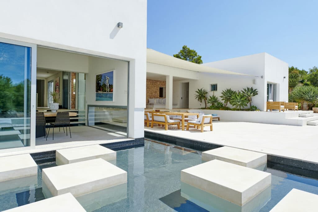 This modern house offers a luxury travel experience with its white walls, large glass doors, and an outdoor patio. The patio features a table with chairs, surrounded by greenery. A small reflective pool with stepping stones leads to the entrance, under a clear blue sky. David Pearson Travel