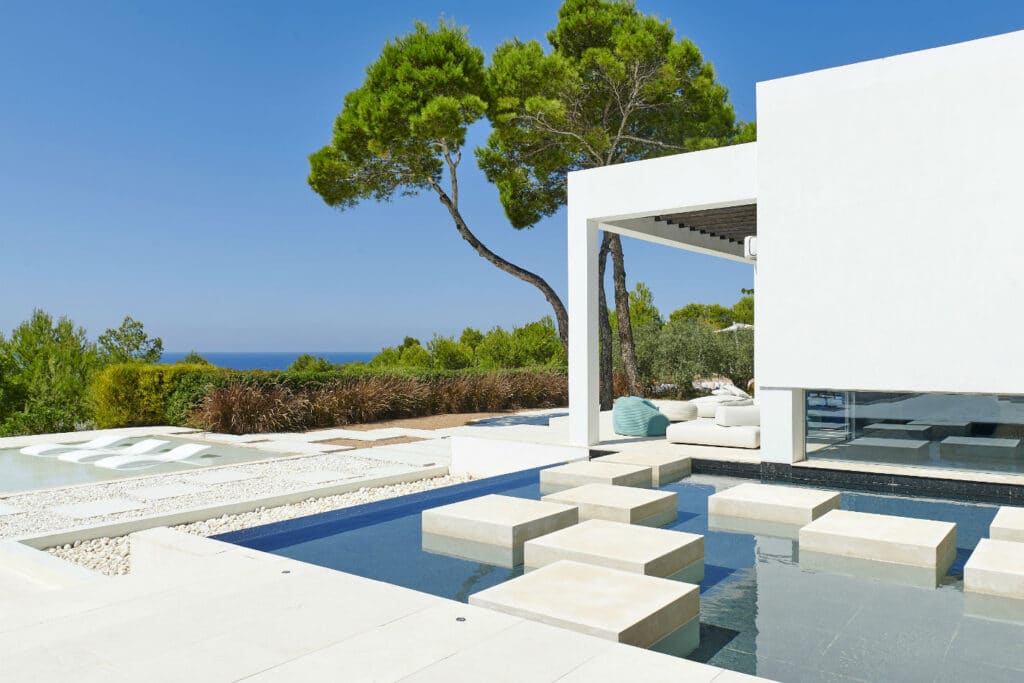 Modern white villa with an open living space overlooking a pool with large stone steps. Surrounded by lush greenery and a clear blue sky, this luxury travel gem also offers a glimpse of the sea in the distance. David Pearson Travel