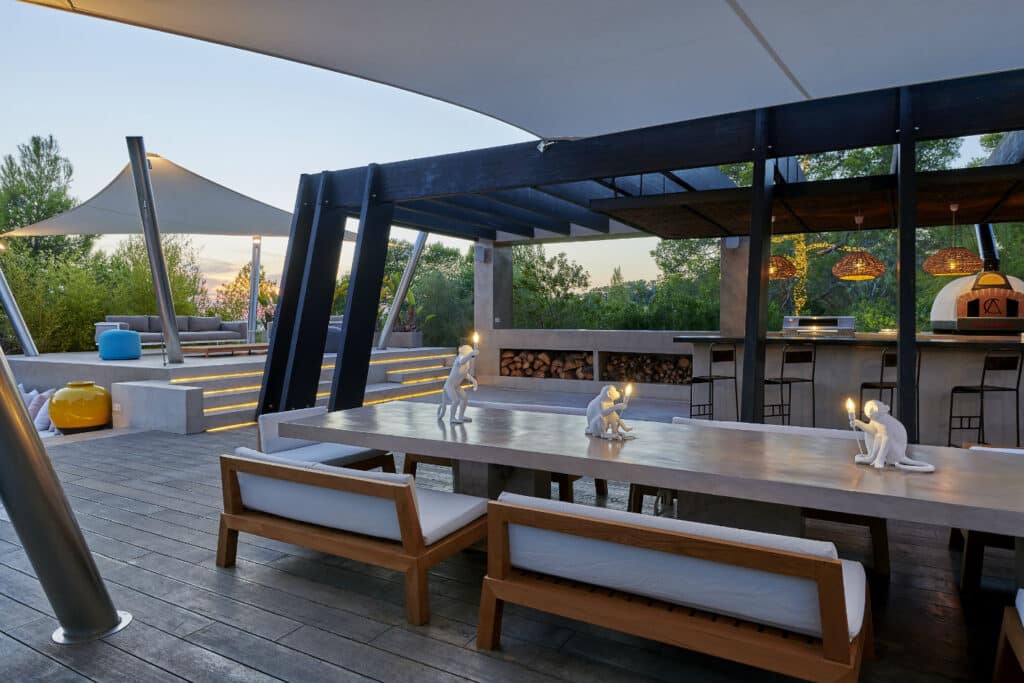 Modern outdoor patio with a long dining table and wooden benches offers a taste of luxury travel. Decorative monkey lamps adorn the table, elevating the ambiance. A bar area with stools is set in the background, surrounded by greenery and shaded by elegant canopies. David Pearson Travel