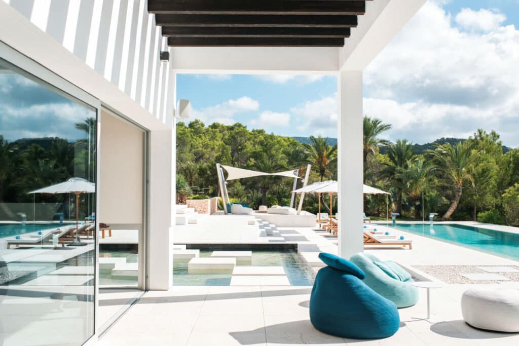 Experience luxury travel at its finest with a modern outdoor patio featuring a pool, shaded lounge chairs, and bean bag seating. Surrounded by lush greenery and palm trees under a partly cloudy sky, this contemporary architectural design boasts a spacious, open layout perfect for relaxation. David Pearson Travel