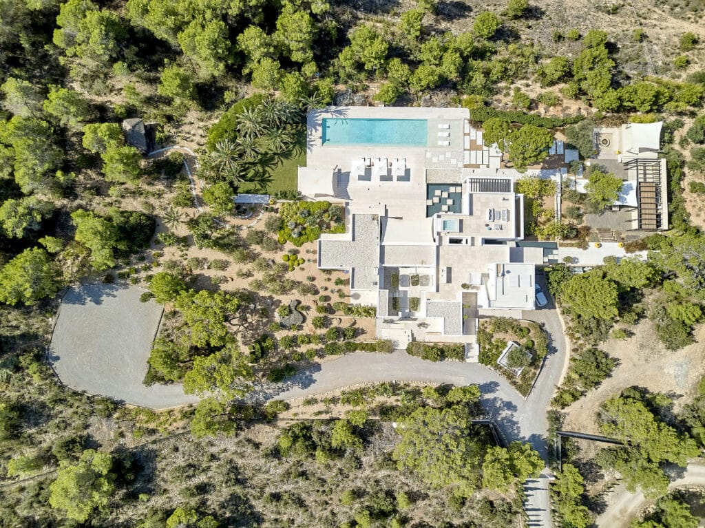 Aerial view of a modern, luxurious villa perfect for luxury travel, surrounded by trees. The property includes a large swimming pool, multiple terraces, and landscaped gardens. A winding driveway leads to the house, set amidst a natural, forested environment. David Pearson Travel