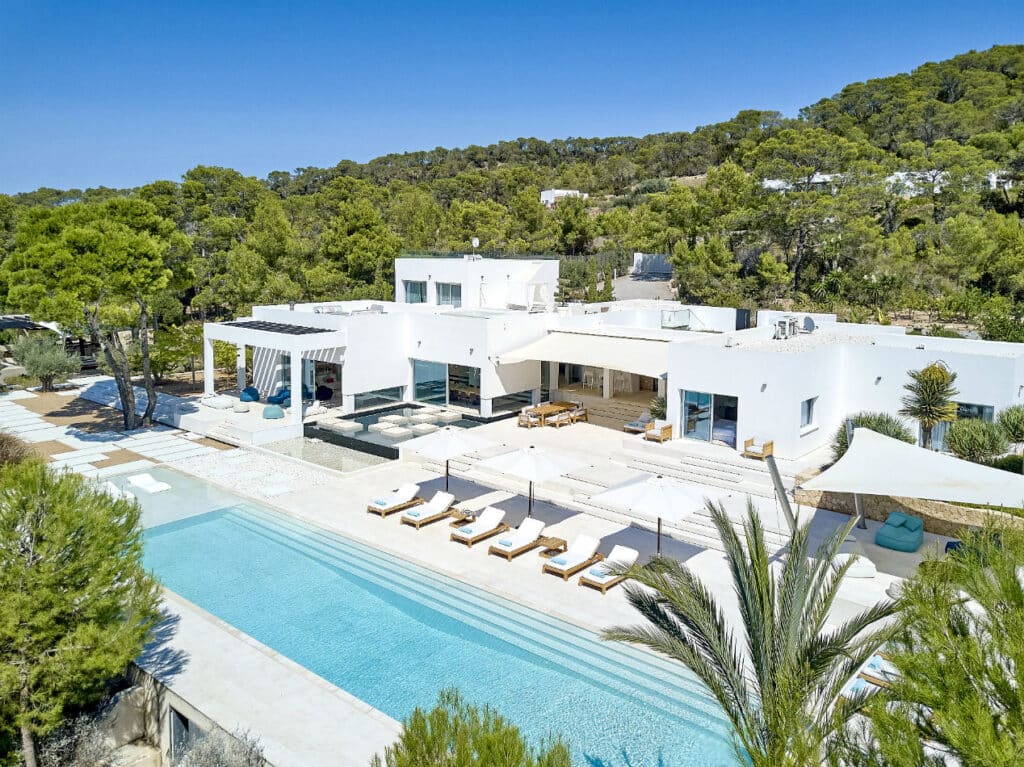 A luxurious modern villa ideal for luxury travel enthusiasts, featuring a large rectangular pool surrounded by sun loungers. The white building is set amidst lush greenery and trees under a clear blue sky, creating a serene and upscale atmosphere. David Pearson Travel