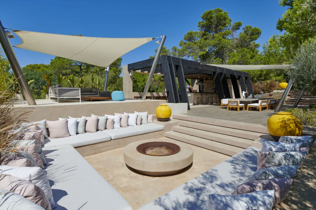 Experience the epitome of luxury travel with an outdoor lounge featuring a sunken seating area around a central fire pit, enveloped by lush greenery. A shaded daybed and covered dining area with wooden furniture create the perfect escape, while bright yellow planters add a vibrant touch. David Pearson Travel