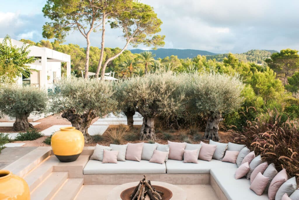 Outdoor patio with a sunken seating area, featuring white cushions and numerous throw pillows. Surrounded by olive trees and greenery, with a view of distant hills that exudes luxury travel vibes. A round fire pit and large yellow vases add decorative elements. David Pearson Travel