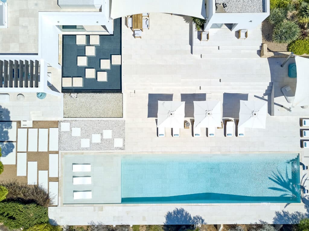 Aerial view of a modern, minimalist outdoor space with a rectangular swimming pool, lined with white sun loungers and umbrellas. Geometric stone pathways and decorative pebbled areas surround the pool, enhancing the luxury travel vibe of this sleek design. David Pearson Travel