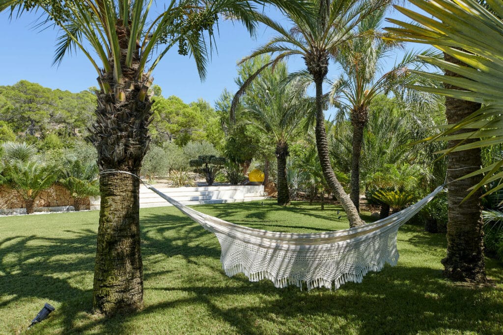 A white fringed hammock, epitomizing luxury travel, is tied between two palm trees on a grassy lawn. Around it flourish tropical plants and trees, with a clear blue sky and distant greenery painting a serene outdoor setting. David Pearson Travel
