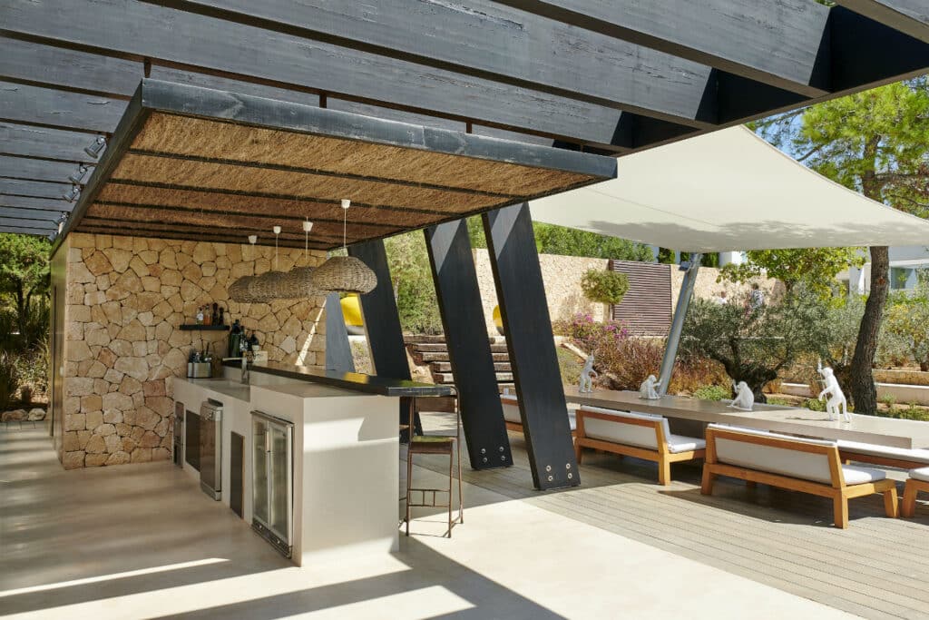 Outdoor kitchen with a stone backsplash, modern countertops, and bar stools. A shaded seating area with cushioned benches and a sail canopy evokes the ambiance of luxury travel, surrounded by lush greenery and a sunny garden. David Pearson Travel