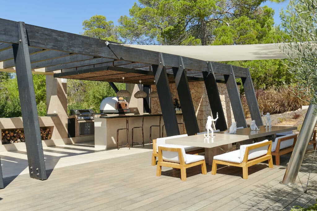 This luxury outdoor kitchen, inspired by upscale travel experiences, showcases a modern pergola with a built-in grill, pizza oven, and elegant bar seating. A long dining table with white chairs sits nearby, surrounded by trees and covered by a shade sail for ultimate comfort. David Pearson Travel