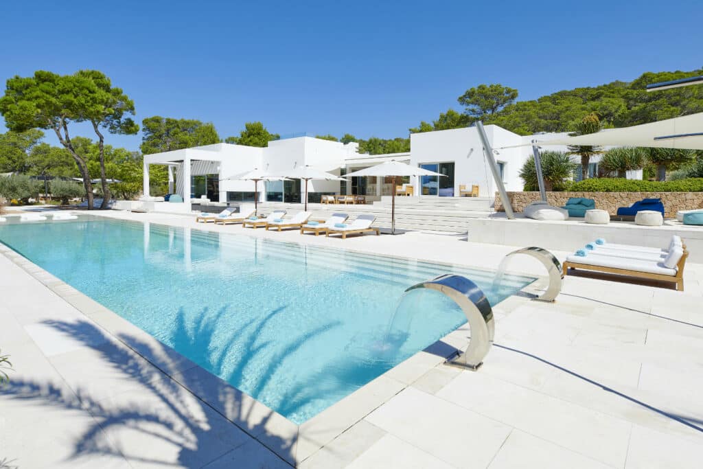 Experience the epitome of luxury travel at this modern villa, featuring a spacious swimming pool with a waterfall. Sun loungers, white umbrellas, and lush greenery create an oasis of relaxation under the clear blue sky. David Pearson Travel