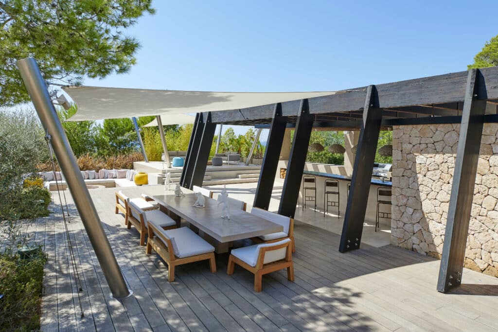 Outdoor dining area exudes luxury travel, featuring a long wooden table and cushioned chairs under a modern pergola. A stone wall graces the right side, with lush greenery in the background and a white sail shade overhead. The sunny setting is serene and inviting. David Pearson Travel