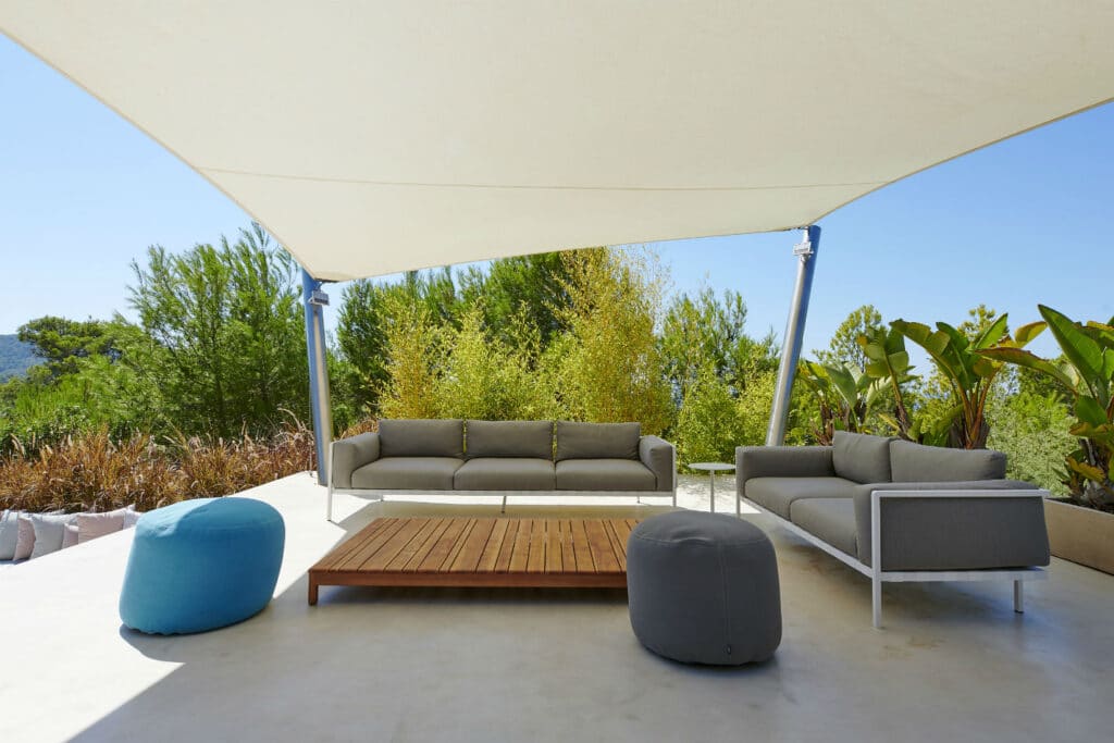 Outdoor patio with two gray sofas and a blue pouf under a large white canopy evokes a luxury travel vibe. A wooden coffee table sits in the center, surrounded by lush green plants, against the backdrop of a clear blue sky. The serene and minimalist setting promises an escape into tranquility. David Pearson Travel