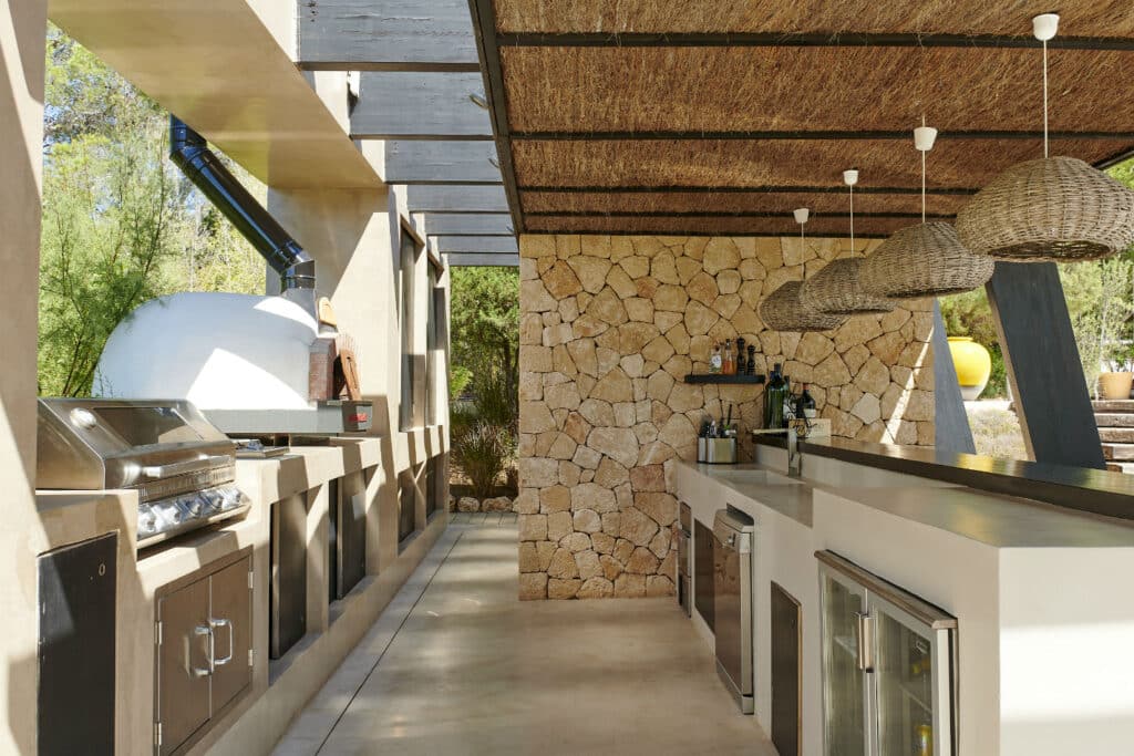 Outdoor kitchen with a stone wall backdrop, a wooden pergola, and modern appliances offers a taste of luxury travel at home. The kitchen features a grill, oven, and countertop with hanging woven pendants. Surrounded by greenery, it provides a stylish and natural ambiance. David Pearson Travel
