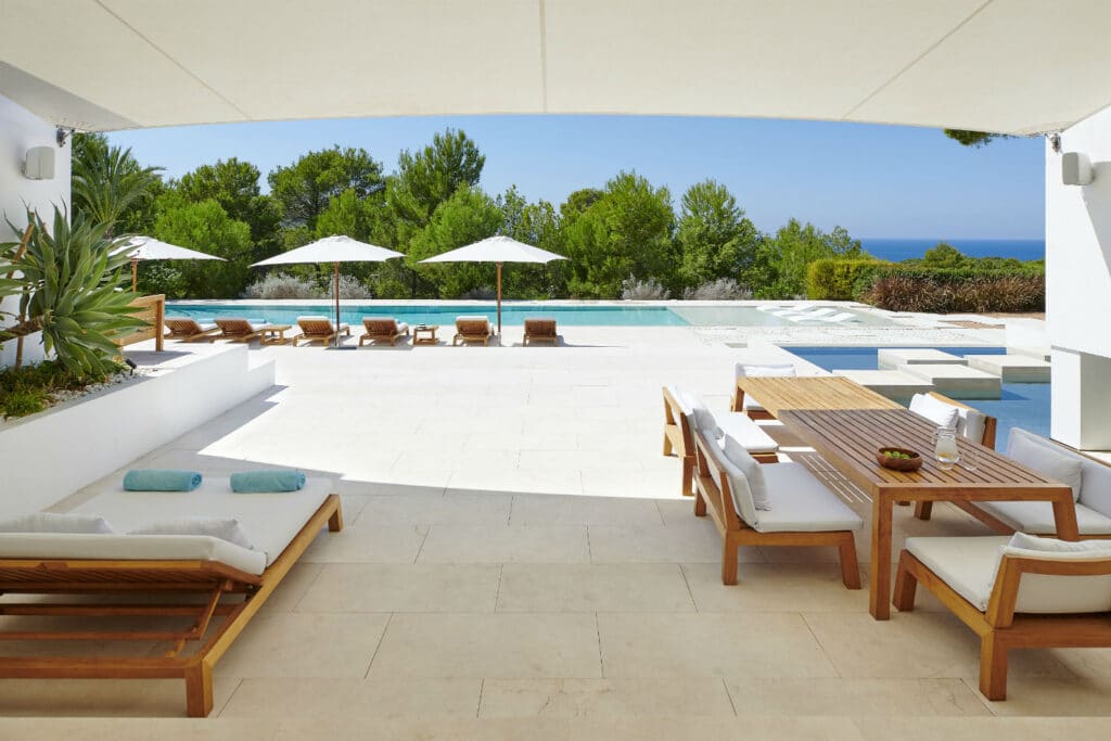 Experience the epitome of luxury travel with a stunning outdoor patio featuring wooden furniture, a sleek dining table, and plush loungers. Unwind by a pristine pool, shaded by umbrellas and embraced by lush greenery, while savoring breathtaking ocean views under a clear blue sky. David Pearson Travel