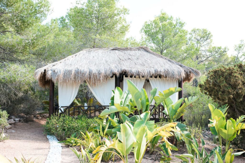 A thatched-roof gazebo with white curtains stands in a garden surrounded by lush greenery and tropical plants, epitomizing luxury travel. Trees fill the background under a bright sky, creating an idyllic retreat. David Pearson Travel