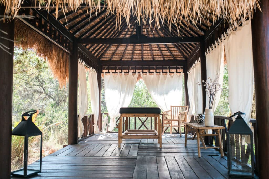 A rustic outdoor gazebo with a wooden floor and thatched roof exudes luxury travel vibes, featuring white drapes tied to its sides. Inside, a massage table and two wooden chairs are elegantly arranged under decorative lanterns, with lush trees and greenery as the perfect backdrop. David Pearson Travel