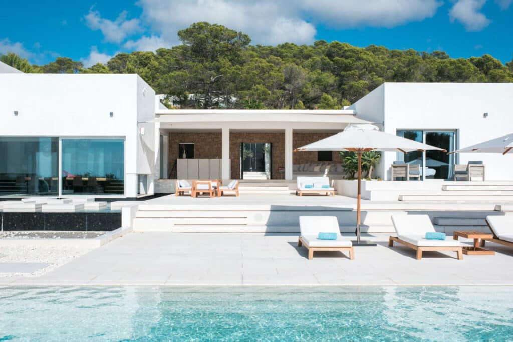 Experience luxury travel at its finest in this modern white villa, boasting large glass windows and a spacious patio with elegant lounge chairs and umbrellas. A clear swimming pool graces the foreground, framed by lush green trees and a vivid blue sky completing the serene escape. David Pearson Travel