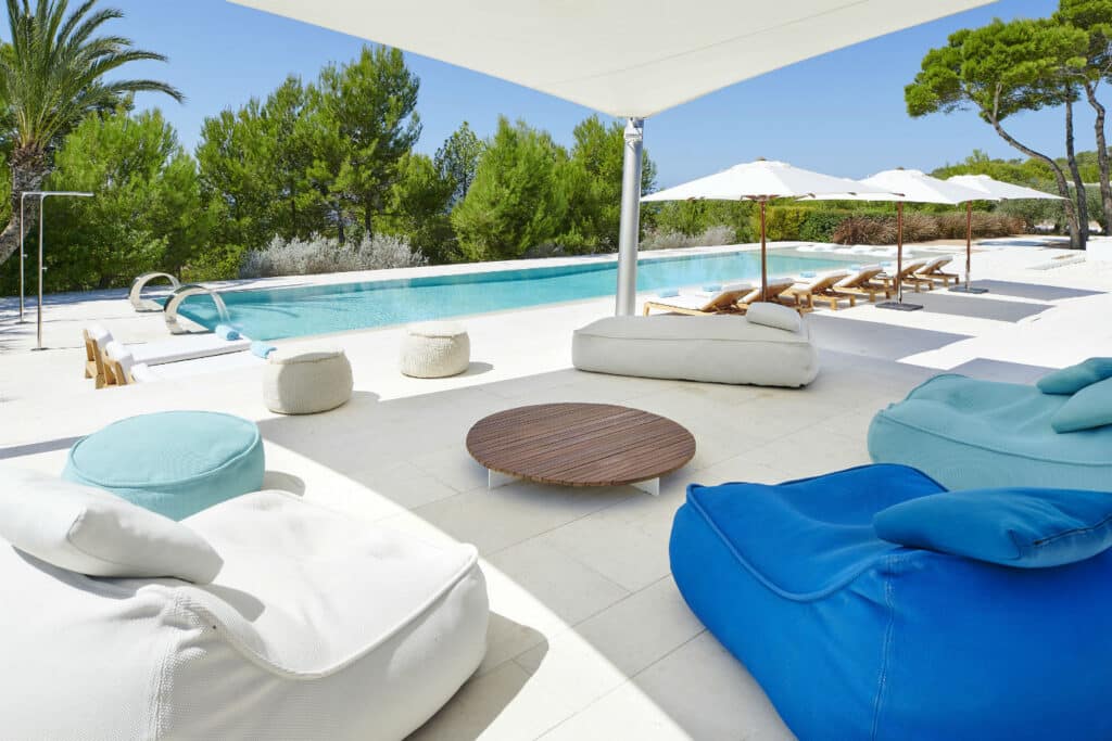 A luxurious outdoor area perfect for luxury travel enthusiasts, with a pool surrounded by trees. The patio features modern bean bag chairs and a round wooden table. Sun loungers with umbrellas are lined up near the pool on the bright tiled floor, ready for ultimate relaxation. David Pearson Travel