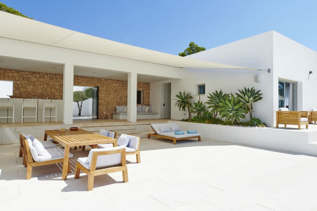 Modern white villa with clean lines, perfect for luxury travel enthusiasts. Featuring a spacious patio with wooden outdoor furniture, cushioned lounge chairs, and a table. Large potted plants add greenery, while stone wall accents and clear blue sky complete the serene setting. David Pearson Travel