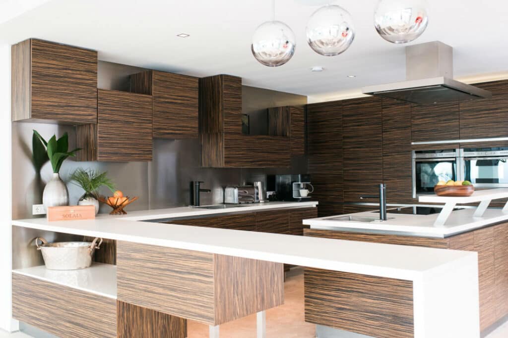 Experience a kitchen that feels like luxury travel at home, with wood-paneled cabinets, white countertops, and stainless steel appliances. Under the glow of three spherical pendant lights, a plant, decorative bowl, and wooden box adorn the counter while a metal bowl rests on a lower shelf. David Pearson Travel