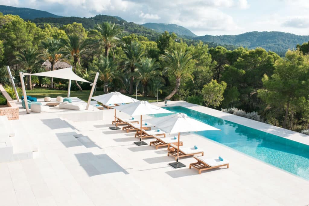 Experience the essence of luxury travel in this opulent outdoor oasis. Lounge chairs with white umbrellas are perfectly placed beside an infinity pool, surrounded by palm trees and lush greenery. Enjoy breathtaking mountain views and a chic shaded seating area, epitomizing ultimate relaxation. David Pearson Travel