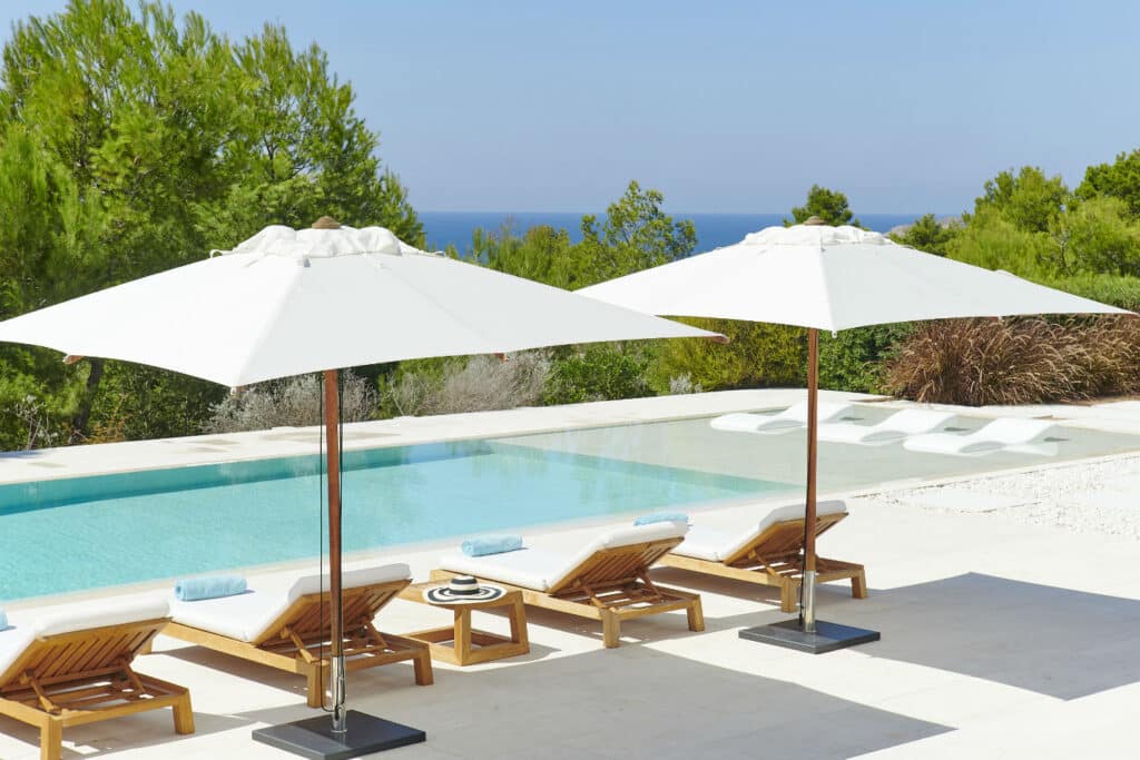 Experience the epitome of luxury travel with a serene view of a lavish outdoor pool area, featuring two wooden lounge chairs under white umbrellas. The pool overlooks lush green trees and a clear blue sky, with the ocean visible in the background. David Pearson Travel