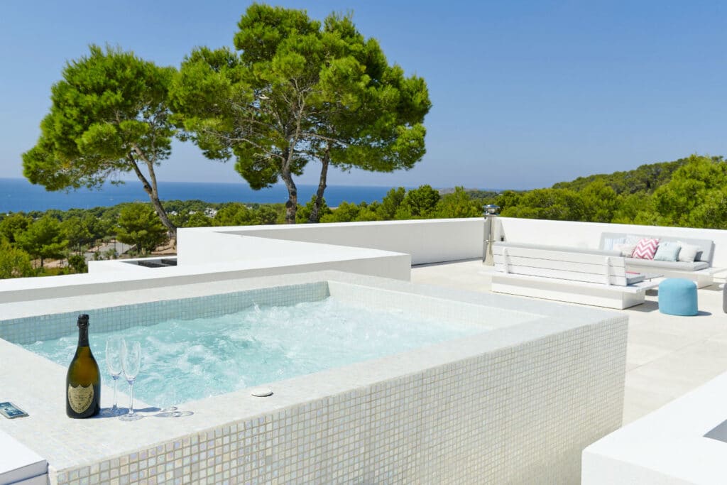 Experience the essence of luxury travel on this modern rooftop terrace. Relax in the jacuzzi with a champagne toast, overlooking lush greenery and the sea. A white sofa and blue pouf invite you under a clear blue sky for an unforgettable retreat. David Pearson Travel