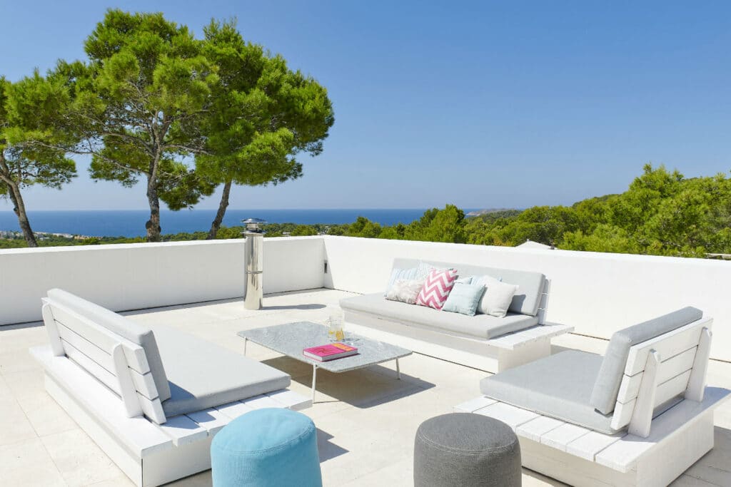 A modern outdoor patio with white and gray seating is set against a backdrop of lush green trees and a clear blue sky, epitomizing the essence of luxury travel. The sea shimmers in the distance, while a glass-top table holds a tray, a book, and brightly colored cushions for accents. David Pearson Travel