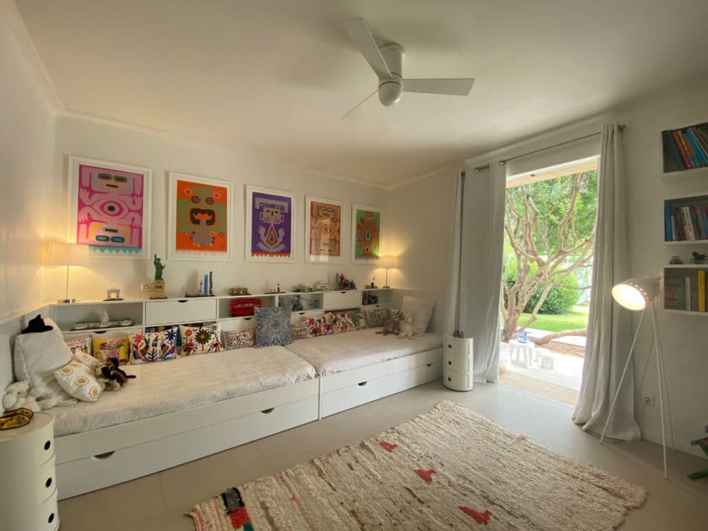 A cozy childrens bedroom with a daybed under colorful framed art exudes the charm of luxury travel. Shelving displays toys and books, while a large window overlooks a green garden. A standing lamp and cushioned rug complete the space, with a ceiling fan above, creating a soothing retreat. David Pearson Travel