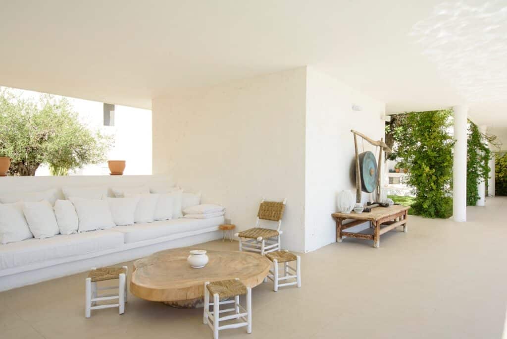 A minimalist outdoor seating area exudes luxury travel vibes with a long white sofa adorned with plush pillows, a round wooden coffee table, and small white stools. A rustic bench holds a statement gong, while lush greenery and an olive tree infuse freshness into the serene space. David Pearson Travel