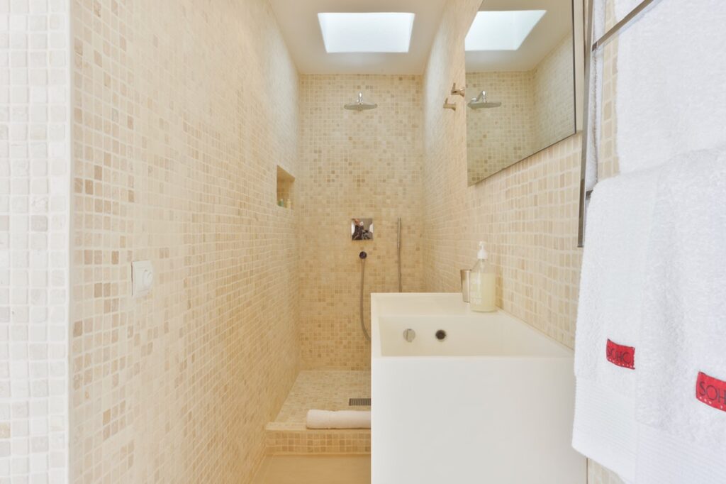 Minimalist beige-tiled bathroom reminiscent of luxury travel suites, featuring a walk-in shower with a rain showerhead. A built-in shelf, rectangular mirror, and white sink with dispenser enhance the elegance. Towels hang neatly on a rack as soft natural light filters through a skylight. David Pearson Travel