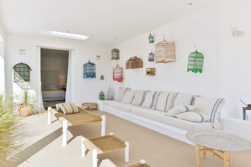 A white patio with a long cushioned bench and several pillows exudes a luxury travel vibe. Colorful birdcages decorate the white wall above. Two woven benches and a round table sit on the beige floor, while a doorway leads to a bedroom with a visible bed. Bright and airy ambiance. David Pearson Travel