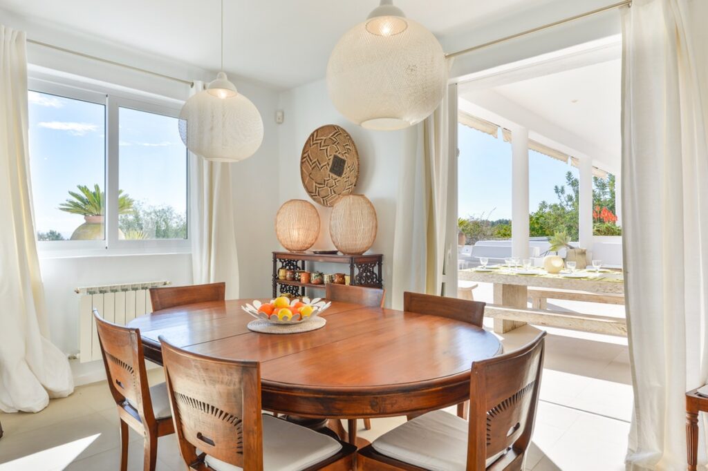 The bright dining area exudes a touch of luxury, with a wooden table set for four beneath elegant pendant lights. Sunlight streams through the window and open sliding doors, seamlessly connecting to an outdoor dining space. Decorative baskets add charm to the side console, reminiscent of luxury travel aesthetics. David Pearson Travel